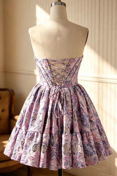 A short off-the-shoulder dress with a floral print in shades of pink, purple, and green, displayed on a mannequin in an indoor setting with warm sunlight and panelled walls.