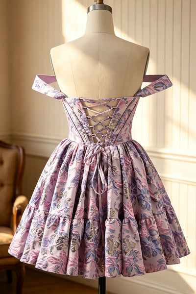 A short off-the-shoulder dress with a floral print in shades of pink, purple, and green, displayed on a mannequin in an indoor setting with warm sunlight and panelled walls.