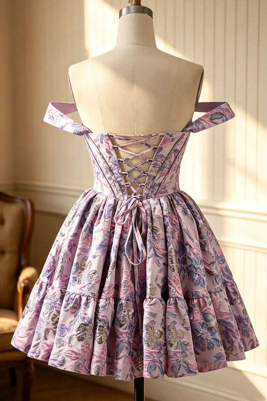 A short off-the-shoulder dress with a floral print in pink, purple, and blue, displayed on a dress form mannequin in a sunlit room.