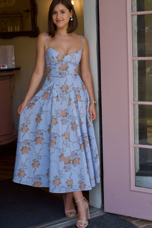 A woman wearing a light blue midi dress with a floral print, standing in a doorway next to a pink door frame.