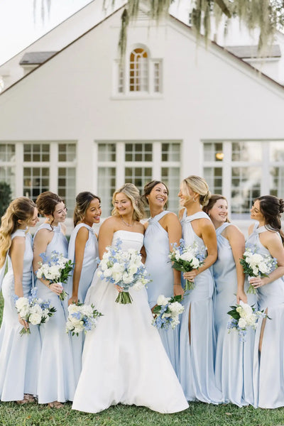 A bride in a white strapless gown stands outdoors with seven bridesmaids wearing matching light blue sleeveless dresses, holding white and blue bouquets in front of a white house.