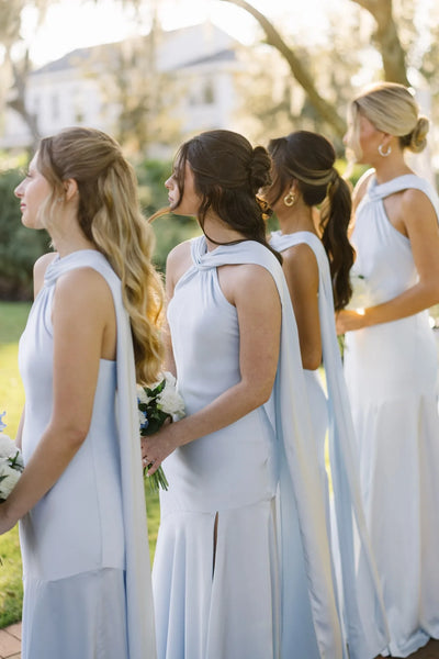 A bride in a white strapless gown stands outdoors with seven bridesmaids wearing matching light blue sleeveless dresses, holding white and blue bouquets in front of a white house.