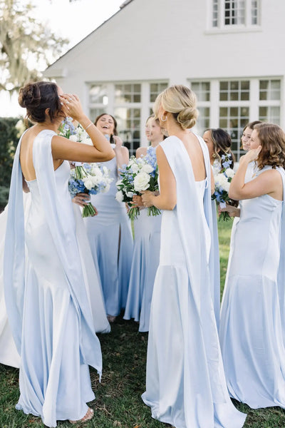 A bride in a white strapless gown stands outdoors with seven bridesmaids wearing matching light blue sleeveless dresses, holding white and blue bouquets in front of a white house.