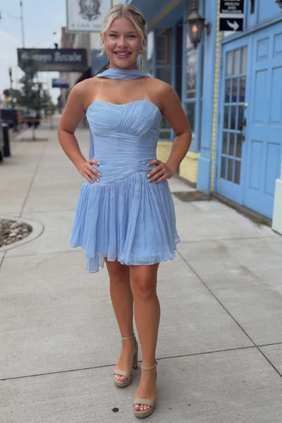 A woman modeling a light blue strapless pleated mini dress with a matching neck scarf, standing on a sidewalk in front of a blue building.