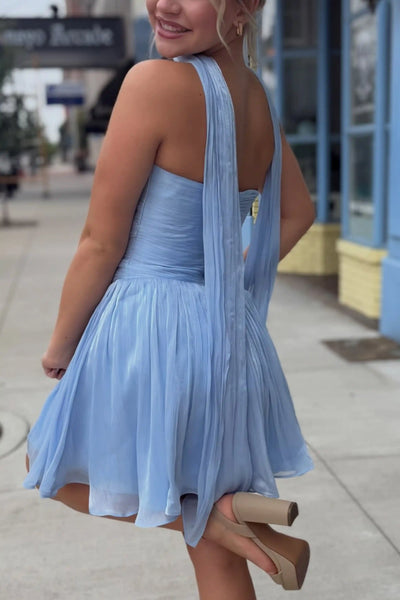 A woman modeling a light blue strapless pleated mini dress with a matching neck scarf, standing on a sidewalk in front of a blue building.