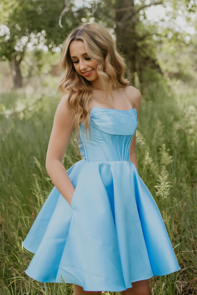 A woman wearing a light blue strapless fit-and-flare dress stands in a grassy outdoor field with trees in the background.
