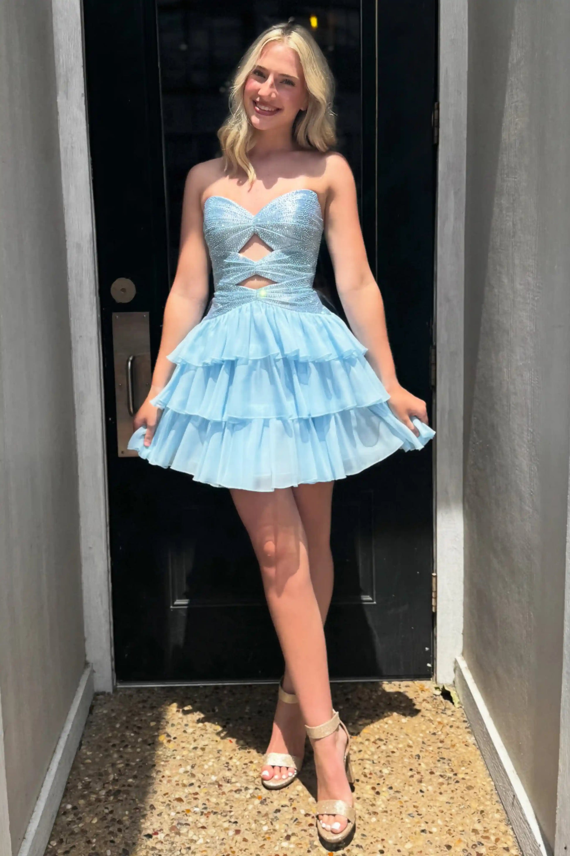 A woman modeling a light blue strapless mini dress with a sweetheart neckline, a cutout at the waist, and a tiered ruffled skirt, standing in a doorway.