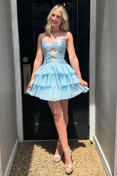 A woman modeling a light blue strapless mini dress with a sweetheart neckline, a cutout at the waist, and a tiered ruffled skirt, standing in a doorway.