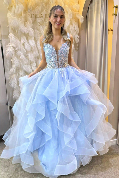 A young woman wearing a light blue ball gown with a beaded bodice and layered ruffle skirt, standing in a fitting room with a feather backdrop.