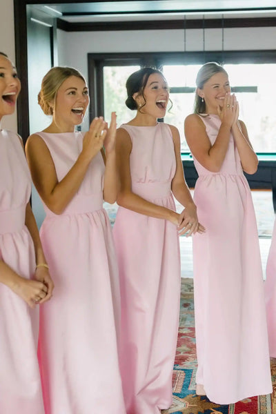Four women wearing sleeveless light pink maxi bridesmaid dresses standing indoors and laughing, with a large window and dark wood trim in the background.