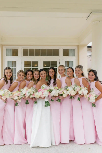 Four women wearing sleeveless light pink maxi bridesmaid dresses standing indoors and laughing, with a large window and dark wood trim in the background.