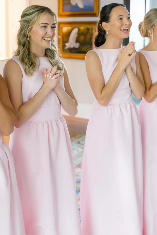 Bridesmaids wearing matching sleeveless light pink A-line dresses, standing indoors and smiling with hands clasped together.