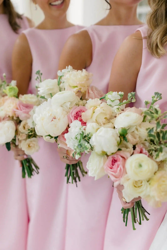 Three bridesmaids in matching light pink sleeveless dresses holding bouquets of white and pink flowers, photographed in a bright indoor setting.