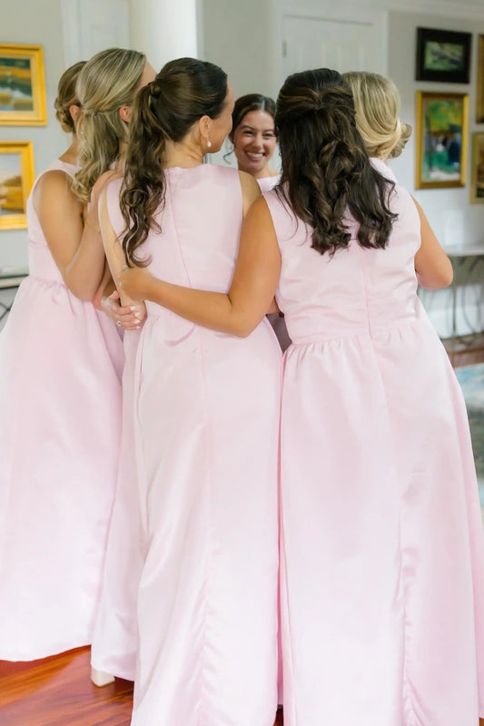Four women wearing matching sleeveless light pink A-line dresses standing together in a bright indoor room.