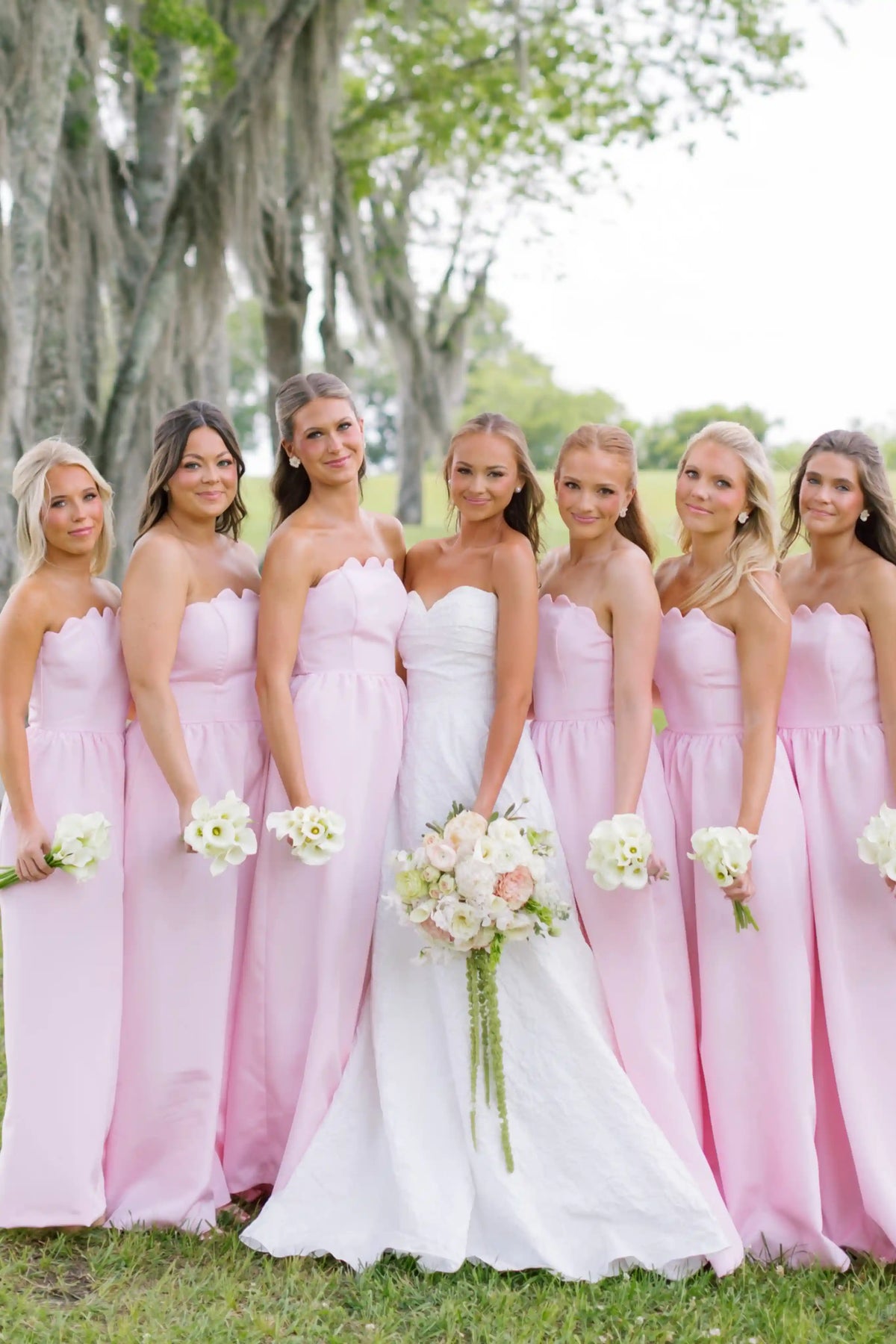 A bride in a white strapless gown stands with five bridesmaids wearing matching light pink strapless dresses, holding flower bouquets outdoors under a tree.