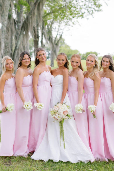 A bride in a white strapless gown stands with five bridesmaids wearing matching light pink strapless dresses, holding flower bouquets outdoors under a tree.