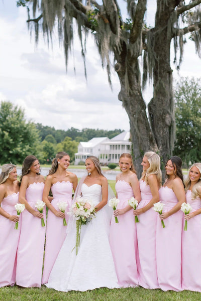 A bride in a white strapless gown stands with five bridesmaids wearing matching light pink strapless dresses, holding flower bouquets outdoors under a tree.