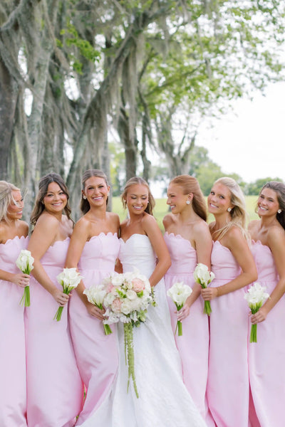 A bride in a white strapless gown stands with five bridesmaids wearing matching light pink strapless dresses, holding flower bouquets outdoors under a tree.