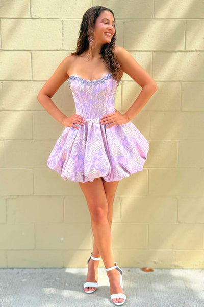 A woman modeling a strapless homecoming mini dress with a corset-style bodice and flared skirt, standing against a beige brick wall.