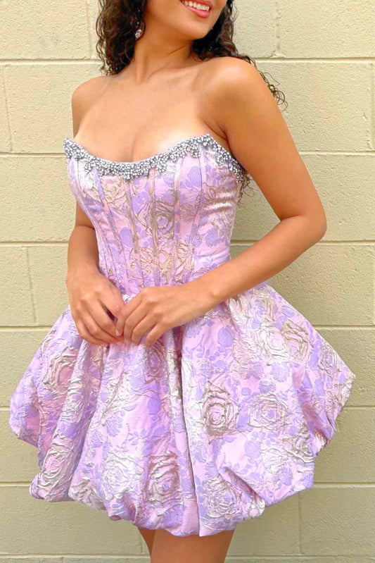 A woman wearing a strapless homecoming short dress with a corseted bodice and flared skirt, standing against a beige brick wall.