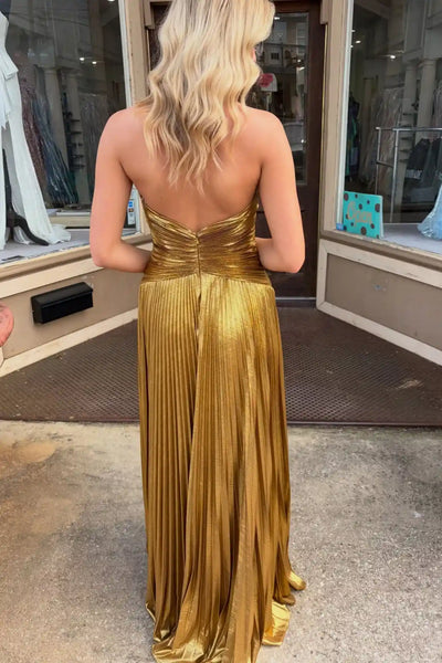 A woman wearing a shimmering metallic gold halter-neck maxi dress with a deep V-neckline, pleated skirt, and thigh-high slit, standing on a sidewalk in front of a storefront.