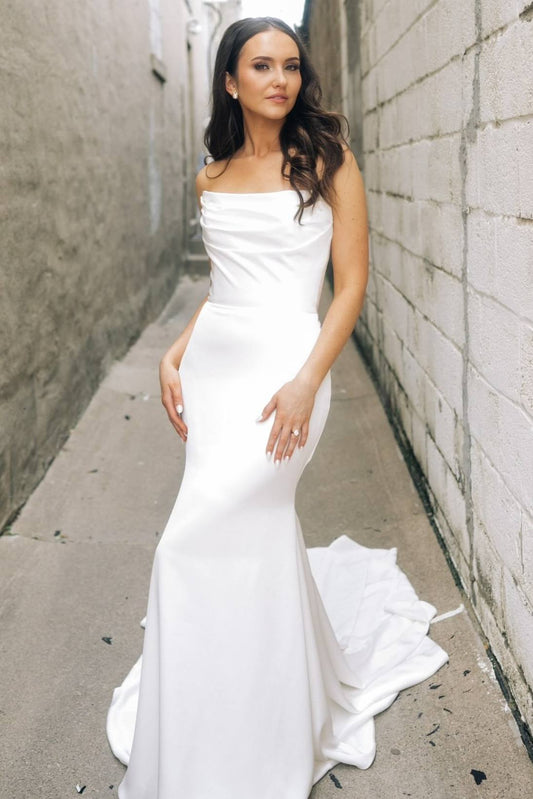 A woman wearing a strapless white mermaid-style wedding gown stands in a narrow outdoor alleyway between textured walls, with the dress train pooling slightly on the ground.