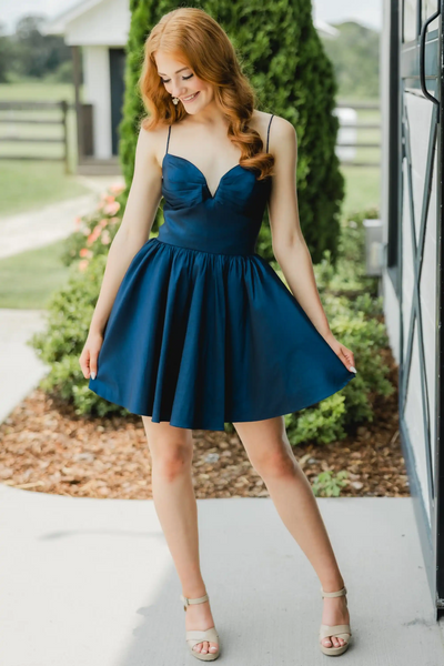 A red-haired woman wearing a short navy blue satin mini dress with spaghetti straps and a flared skirt, posing outdoors in front of a house and greenery.