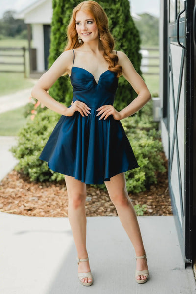 A red-haired woman wearing a short navy blue satin mini dress with spaghetti straps and a flared skirt, posing outdoors in front of a house and greenery.