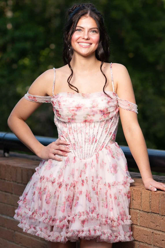 A young woman wearing pink off-the-shoulder tulle dress, posing outdoors against a brick wall and green foliage background.