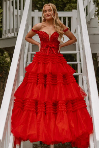 A woman wearing a red off-the-shoulder prom dress with a ruffled tiered skirt, standing on white outdoor stairs.