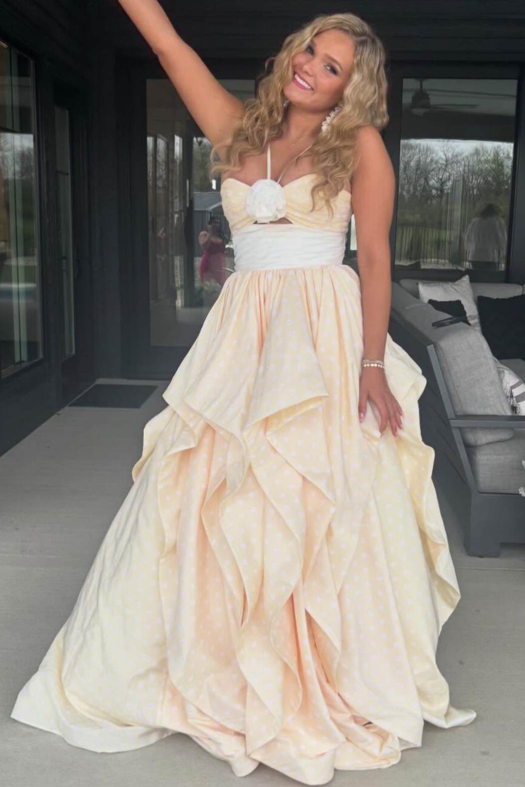 A woman wearing a strapless formal gown with a sweetheart neckline, a white floral bodice detail, a white waistband, and a voluminous layered skirt in a pale peach or cream tone, standing on a patio.