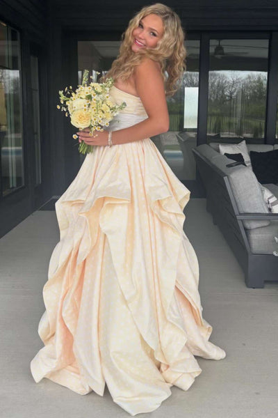 A woman wearing a strapless formal gown with a sweetheart neckline, a white floral bodice detail, a white waistband, and a voluminous layered skirt in a pale peach or cream tone, standing on a patio.