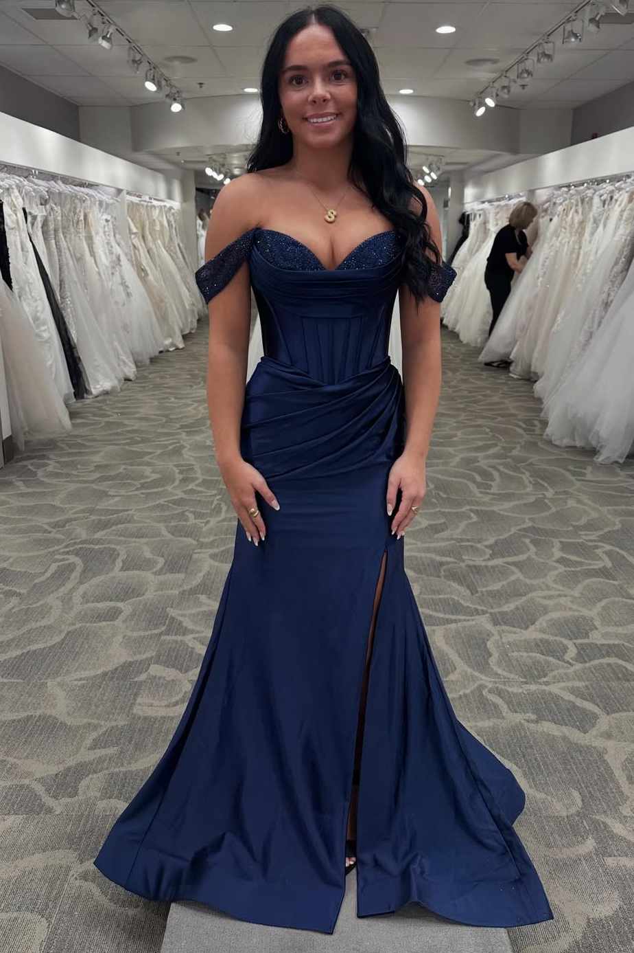 A woman wearing a navy blue off-the-shoulder evening gown with a sweetheart neckline, corset-style bodice, and thigh-high slit, standing in a bridal or formalwear showroom surrounded by white wedding dresses.