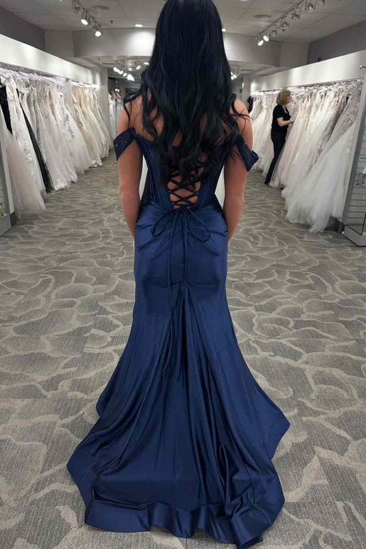 A woman viewed from behind wearing a floor-length royal blue off-the-shoulder evening gown with a lace-up open back, standing in a bridal or formalwear showroom with white dresses hanging in the background.