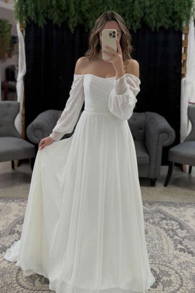 A woman takes a mirror selfie wearing an off-the-shoulder long-sleeve A-line wedding dress in white, standing in a showroom with gray chairs and a black backdrop.
