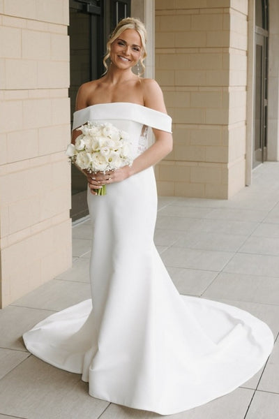 A woman wearing an off-the-shoulder white wedding gown with a slight train, holding a bouquet of white flowers, standing in front of a beige stone building.