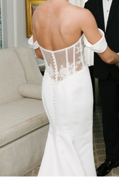 A woman wearing an off-the-shoulder white wedding gown with a slight train, holding a bouquet of white flowers, standing in front of a beige stone building.