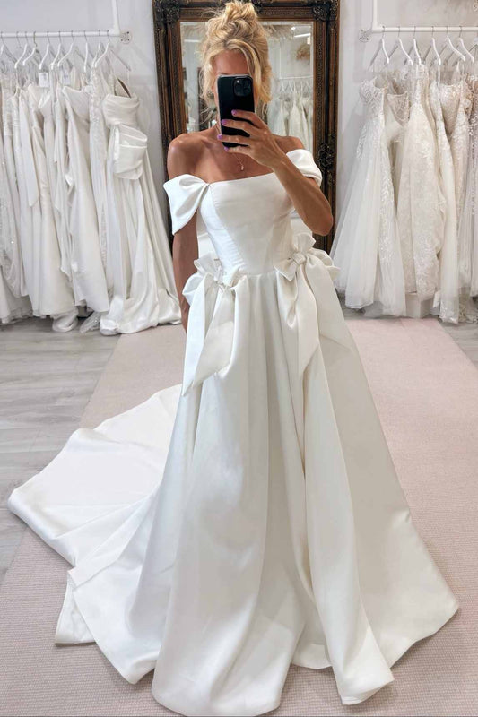 A woman takes a mirror selfie in a bridal shop wearing an off-the-shoulder white satin wedding gown with a structured bodice, large bow details, and a full skirt with a train.