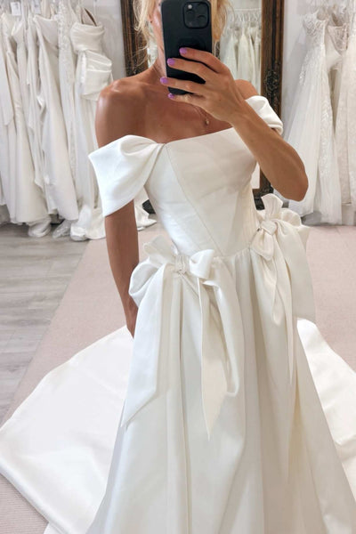A woman takes a mirror selfie in a bridal shop wearing an off-the-shoulder white satin wedding gown with a structured bodice, large bow details, and a full skirt with a train.