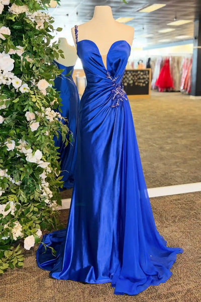 A royal blue floor-length evening gown displayed on a mannequin in an indoor showroom. The dress has a sweetheart neckline, draped bodice, and a slight train, with decorative embellishment at the waist.