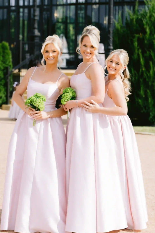 Three blonde bridesmaids wearing matching pale pink sleeveless A-line dresses stand outdoors holding green bouquets in front of a dark fence and greenery.