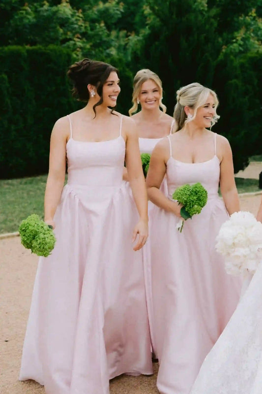 Three bridesmaids wearing matching pale pink sleeveless bridesmaid maxi dresses walk outdoors holding green bouquets, with a bride in a white dress partially visible in the foreground.