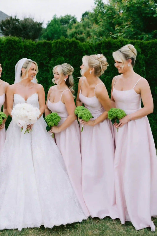 Four bridesmaids in matching light pink sleeveless wedding dresses stand outdoors with green bouquets beside a bride in a white wedding gown, set against a green hedge background.