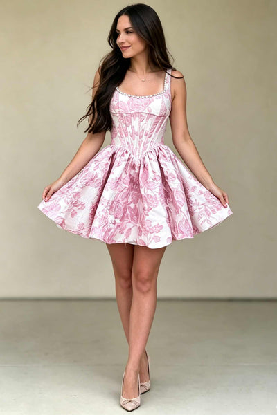 A woman modeling a pink and white Jacquard mini dress with a corset-style bodice and flared skirt, paired with nude heels, against a plain beige studio backdrop.