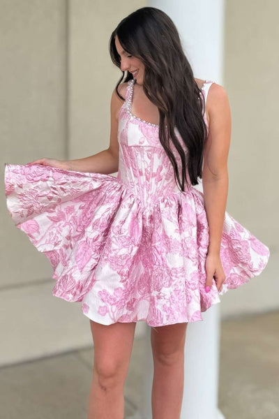 A woman modeling a pink and white Jacquard mini dress with a corset-style bodice and flared skirt, paired with nude heels, against a plain beige studio backdrop.