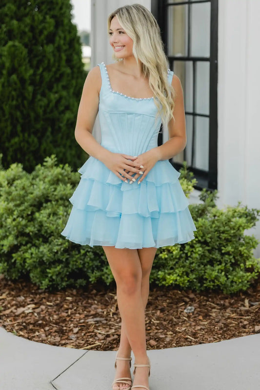A woman wearing a light blue tiered tulle homecoming mini dress with a ruffled sweetheart neckline, standing outdoors in front of a white building with green shrubs.