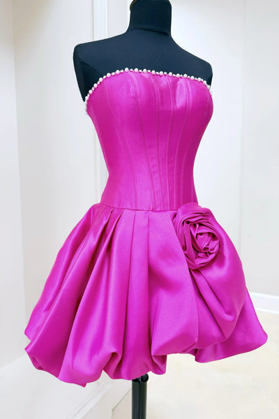 A woman modeling a light blue strapless homecoming mini dress with a structured bodice, flared skirt, and a large fabric rose detail on the side, standing against a white brick wall.