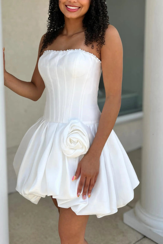 A woman wearing a strapless white corset-style mini dress with a flared skirt and a large fabric rose detail at the waist, standing between white columns.