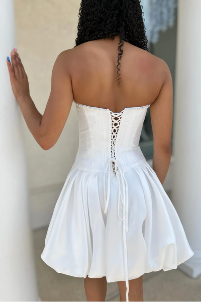 A woman modeling a light blue strapless homecoming mini dress with a structured bodice, flared skirt, and a large fabric rose detail on the side, standing against a white brick wall.