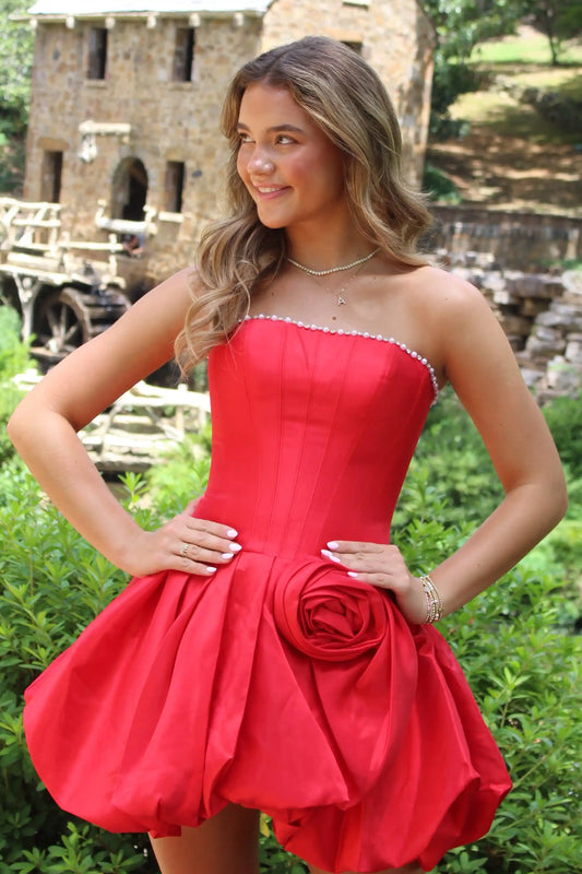 A woman wearing a red strapless homecoming mini dress with a sweetheart neckline, beaded trim, a fitted corset-style bodice, and a large fabric rose detail at the waist, standing outdoors in front of a stone building and greenery.